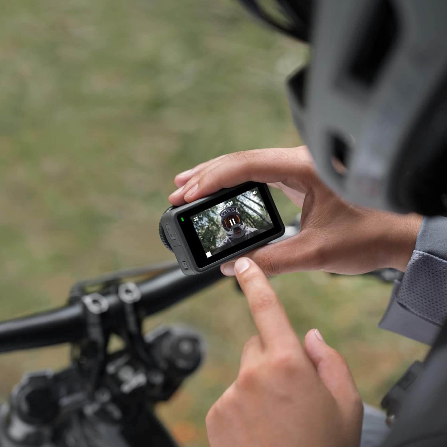 A person wearing biking gear interacts with a DJI Osmo Action 3 camera mounted on a bike handlebar. The image shows the camera’s touchscreen display, which reflects the biker's face with a helmet and goggles.